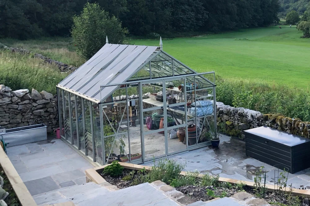 A glass Rhino Greenhouse sits on a stone patio, containing potted plants and gardening supplies. It's surrounded by a lush garden and stone walls, with a green field and forest in the background.