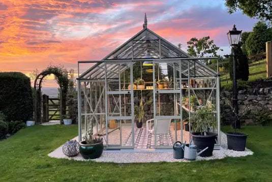 A glass Rhino Greenhouse stands on a gravel area, containing plants and a chair. It is surrounded by a lush garden with trees and a stone wall, under a vibrant sunset sky.