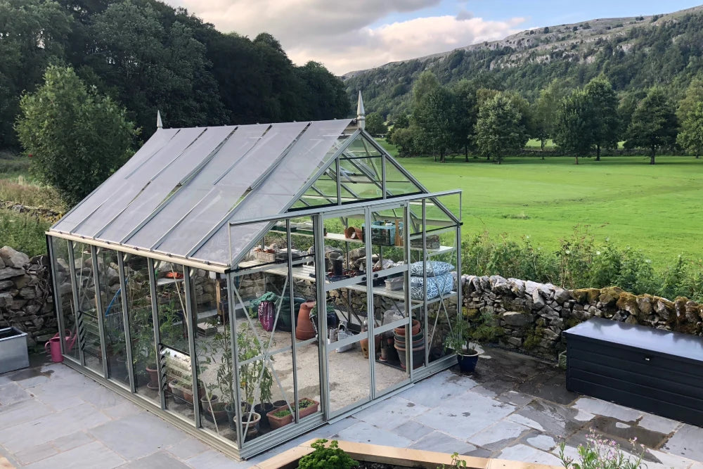A glass Rhino Greenhouse contains various potted plants and gardening tools. It is situated on a stone patio, adjacent to a stone wall, with lush green fields and trees in the background.