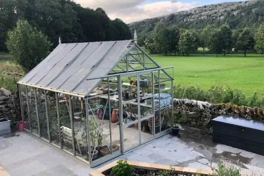 A glass Rhino Greenhouse stands on a stone patio containing various potted plants and gardening tools. It is surrounded by a stone wall and overlooks a grassy field with trees and hills.