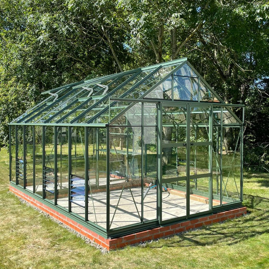 A green metal-framed Rhino Greenhouse stands on a brick foundation, surrounded by lush trees and grass. Sunlight illuminates its glass panels, creating a bright, inviting atmosphere.