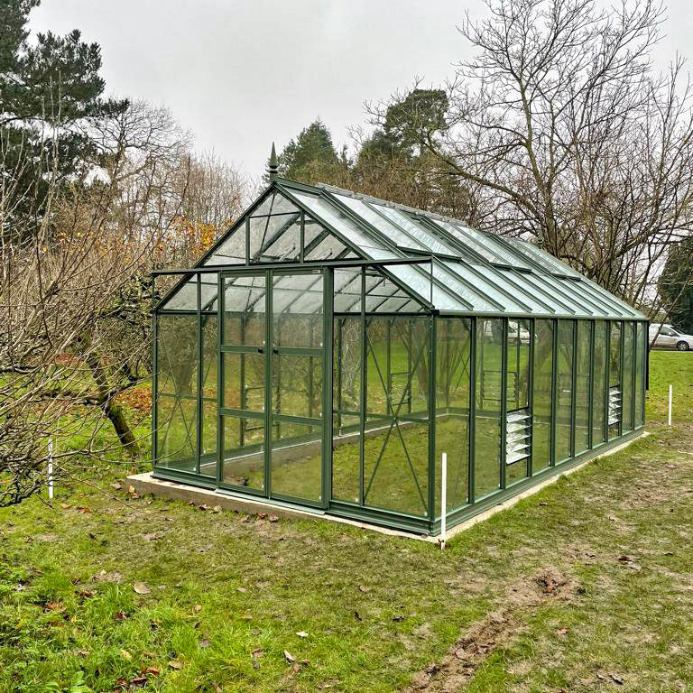 A glass Rhino Greenhouse stands on grass, with trees and overcast sky in the background, displaying modern, green metal framework and clear panels for plant cultivation.