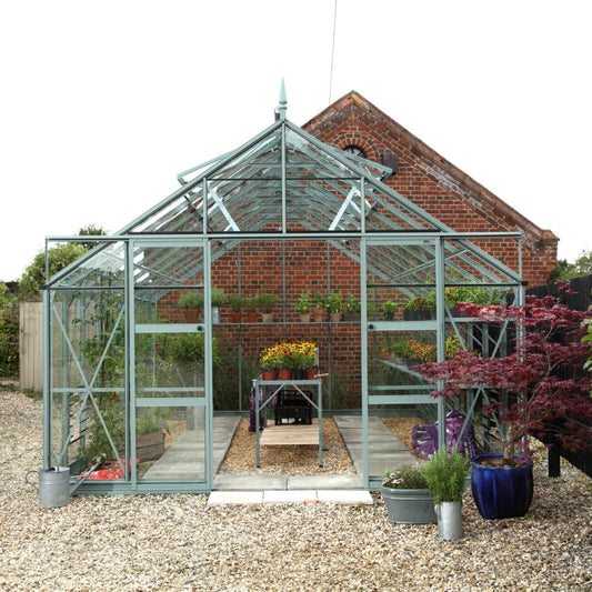 A glass Rhino Greenhouse showcases potted plants and flowers inside, surrounded by a gravel path. It is positioned in front of a brick building, with additional pots on either side.
