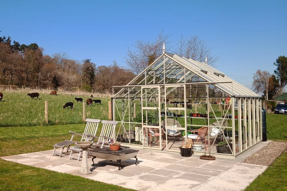 A glass Rhino Greenhouse containing potted plants stands on a patio with chairs and a table. In the background, cows graze in a fenced grassy field under a clear blue sky.