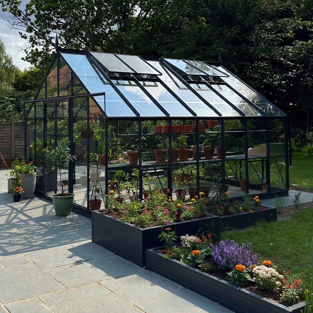 A glass Rhino Greenhouse shelters potted plants, reflecting sunlight, surrounded by blooming flowers in raised beds, with a wooden fence and green trees in the background.