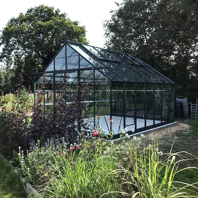A glass Rhino Greenhouse stands in a garden surrounded by lush greenery and various flowering plants under a clear sky.