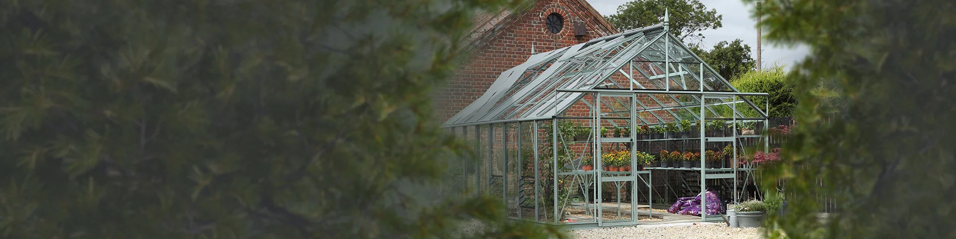 Rhino Greenhouse stands amidst lush foliage, showcasing neatly potted plants inside. Behind, a red brick building with a circular vent creates a rustic garden setting.