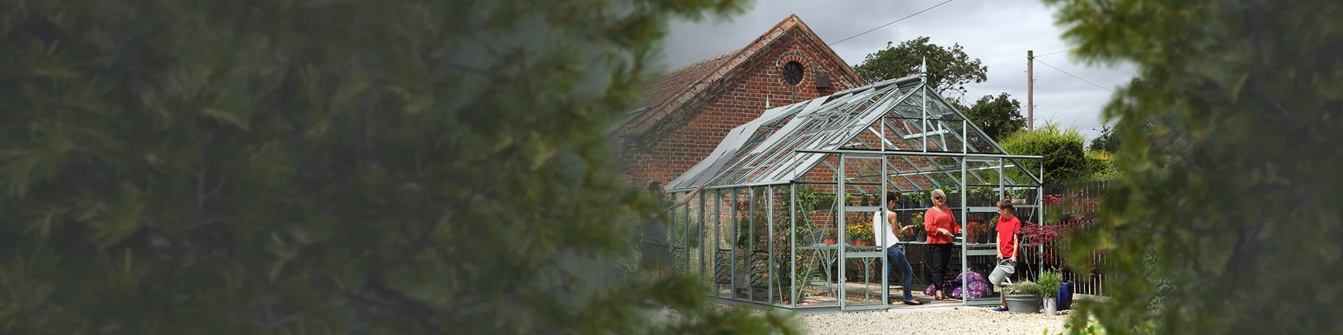 A glass Rhino Greenhouse is in use by three people tending plants inside. It's set in a garden area with trees and a red brick building behind.