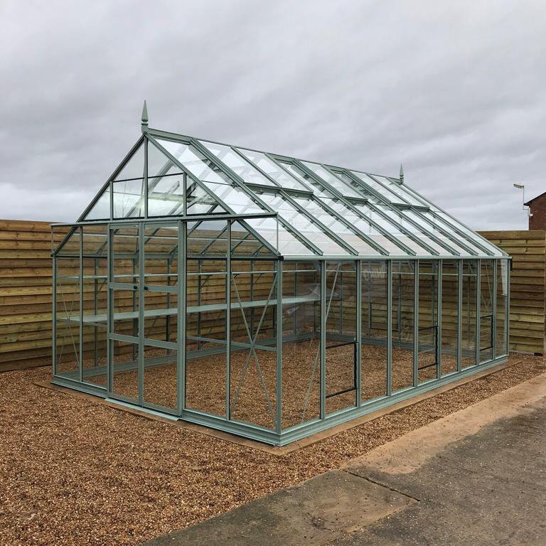 A glass and metal Rhino Greenhouse stands on a gravel surface, surrounded by wooden fencing, under an overcast sky.