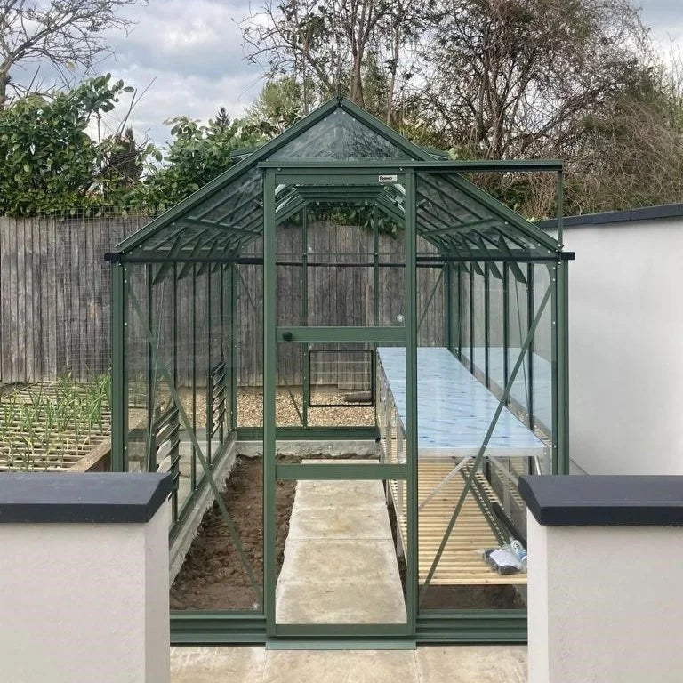 Rhino Greenhouse with a green frame stands open, flanked by shelving inside, set on a concrete path. Surrounding it are trees, a wooden fence, and garden plots.