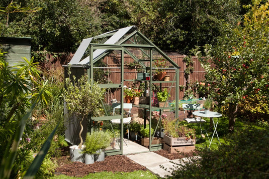 A green Rhino Greenhouse stands in a lush garden, filled with potted plants. Nearby, a small table and chair set complements the greenery. A wooden crate reads, Rhino, Proudly British.