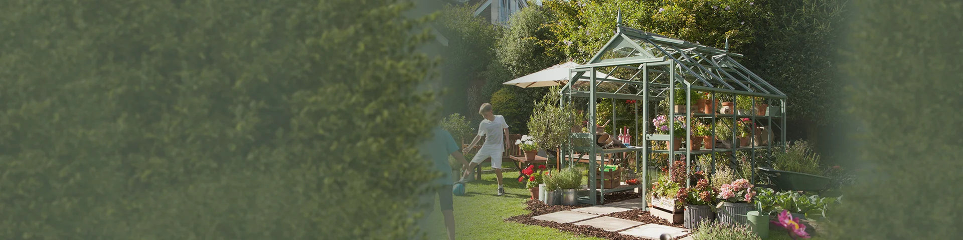 A Rhino Greenhouse filled with potted plants stands in a garden. Nearby, a person walks on the grass under an umbrella and trees, creating a lush outdoor setting.