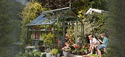 A green Rhino Greenhouse, with potted plants and flowers inside, stands in a lush garden. Nearby, three children interact with the plants, surrounded by leafy trees and garden furniture.