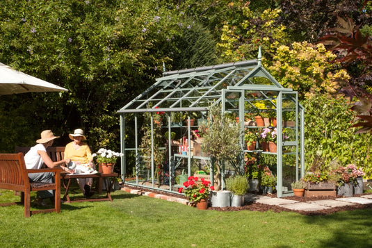 A glass Rhino Greenhouse contains various plants and pots. Nearby, two people sit on a wooden bench under an umbrella, conversing in a lush, sunny garden setting.