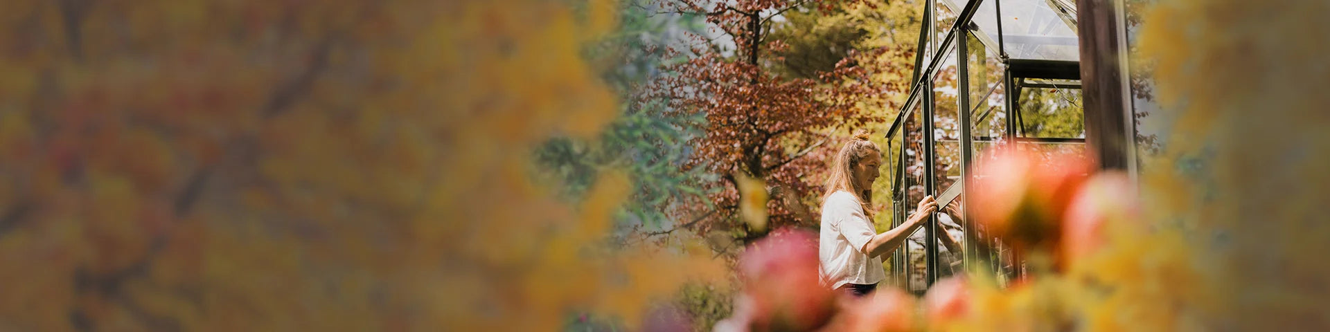 A person tends to a Rhino Greenhouse, adjusting the structure, surrounded by vibrant, blooming flowers and colorful autumn foliage in a serene outdoor garden setting.