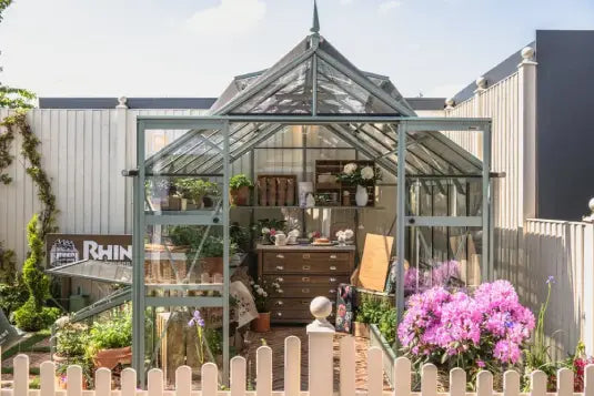 A glass Rhino Greenhouse displays vibrant flowers and gardening tools. It's fenced with a white picket, surrounded by colorful plants. A wooden sign nearby reads Rhino Rhino Greenhouses.