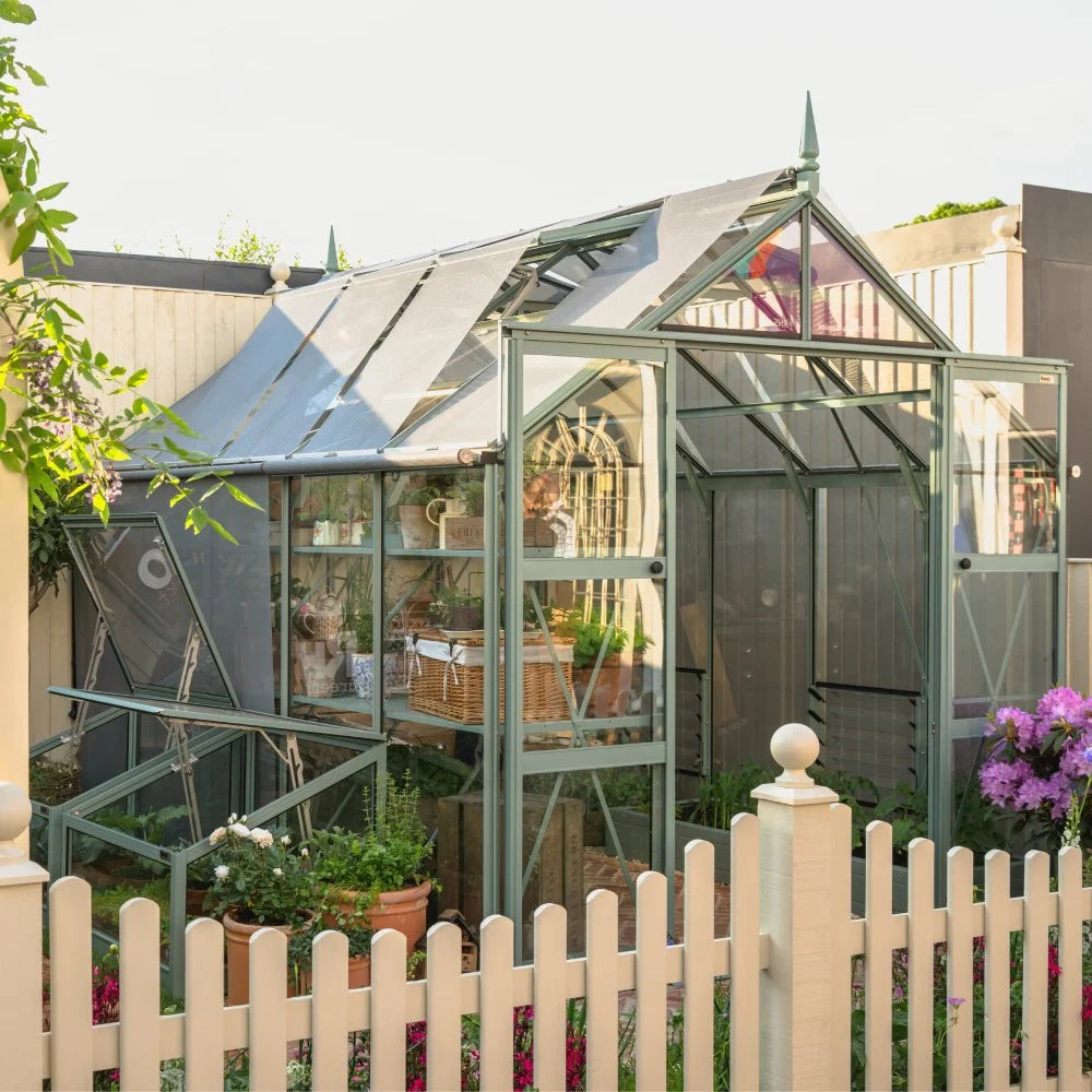 A medium-sized glass Rhino Greenhouse stands in a garden behind a white picket fence, surrounded by potted plants and flowers. It features a sloped roof and houses various gardening tools and plants.