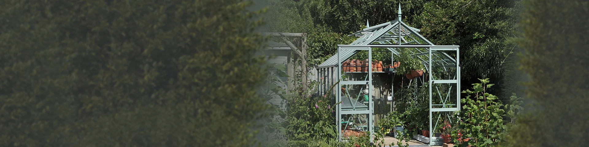 A metal-framed Rhino Greenhouse sits amidst lush greenery, housing potted plants and gardening tools, surrounded by a garden filled with various shrubs and trees.