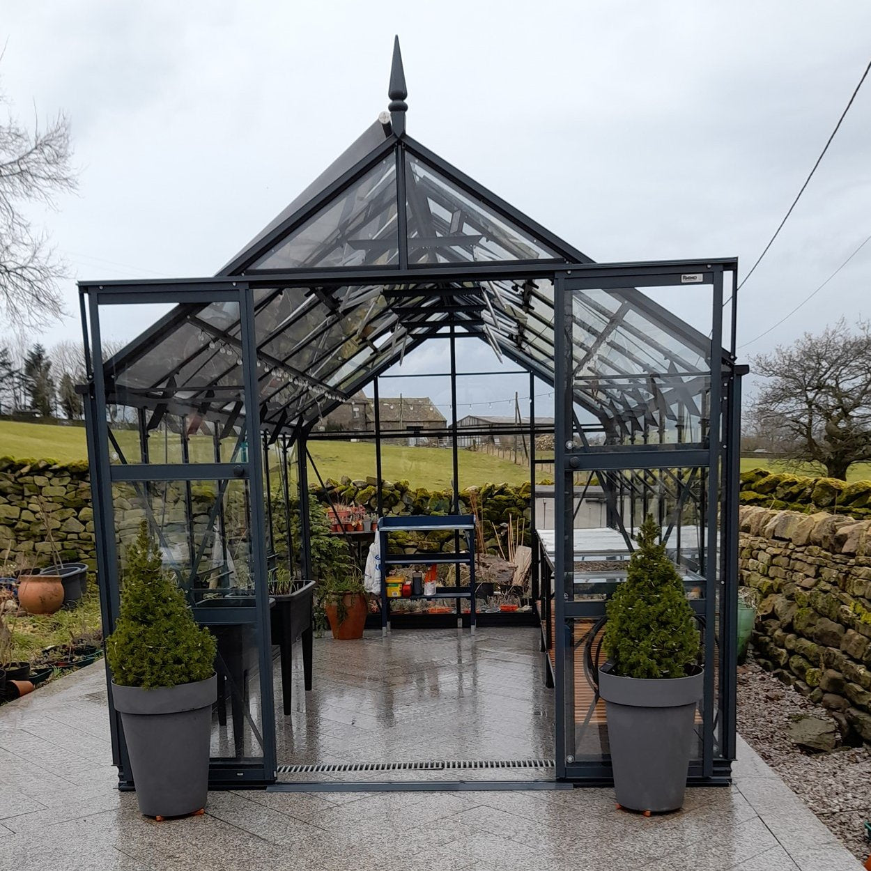 A glass Rhino Greenhouse stands open, revealing potted plants and gardening tools on shelves. It is situated outdoors, surrounded by stone walls and a rural landscape under a cloudy sky.