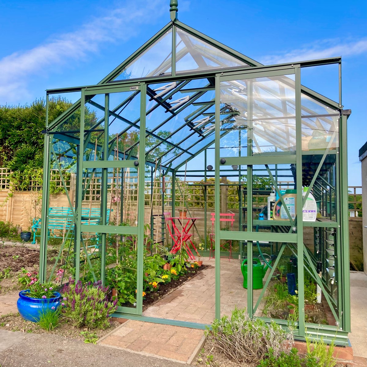 A green Rhino Greenhouse stands with open doors, revealing plants and gardening tools inside. It's surrounded by a garden, with blue sky above. A blue ceramic pot and colorful chairs are visible.