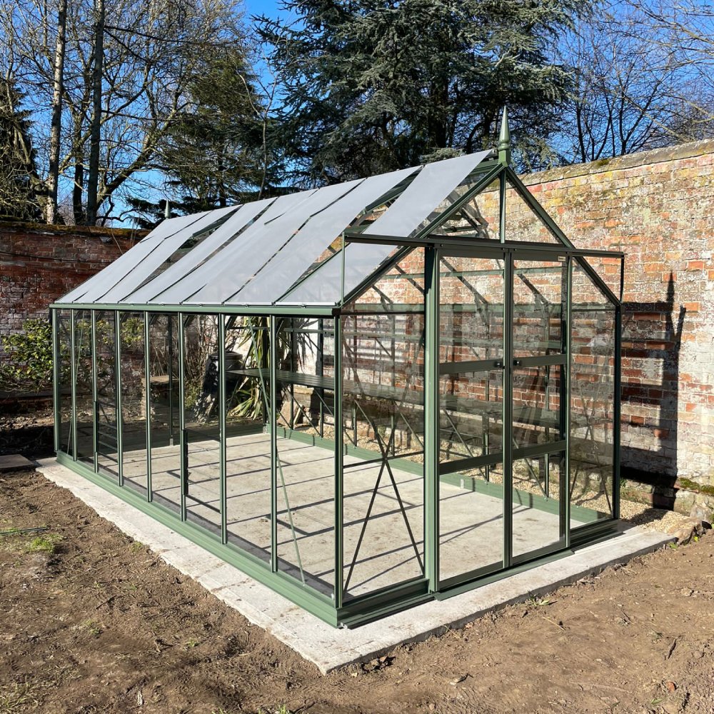 A green, glass Rhino Greenhouse stands on a concrete base. It features a pitched roof and door, situated against a brick wall amidst an outdoor garden setting with trees and sunlight.