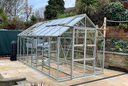 A glass Rhino Greenhouse with a metal frame stands on a patio. It's surrounded by a garden with various plants and a stone retaining wall.