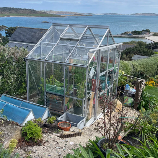 A glass Rhino Greenhouse houses lush plants beside a quaint garden with soil and potted plants. The backdrop features a picturesque ocean view and distant hills under a blue sky.