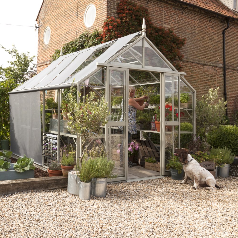 A woman tends to plants inside a glass Rhino Greenhouse. A dog sits outside on a gravel path, next to potted plants. A brick building and lush greenery form the backdrop.