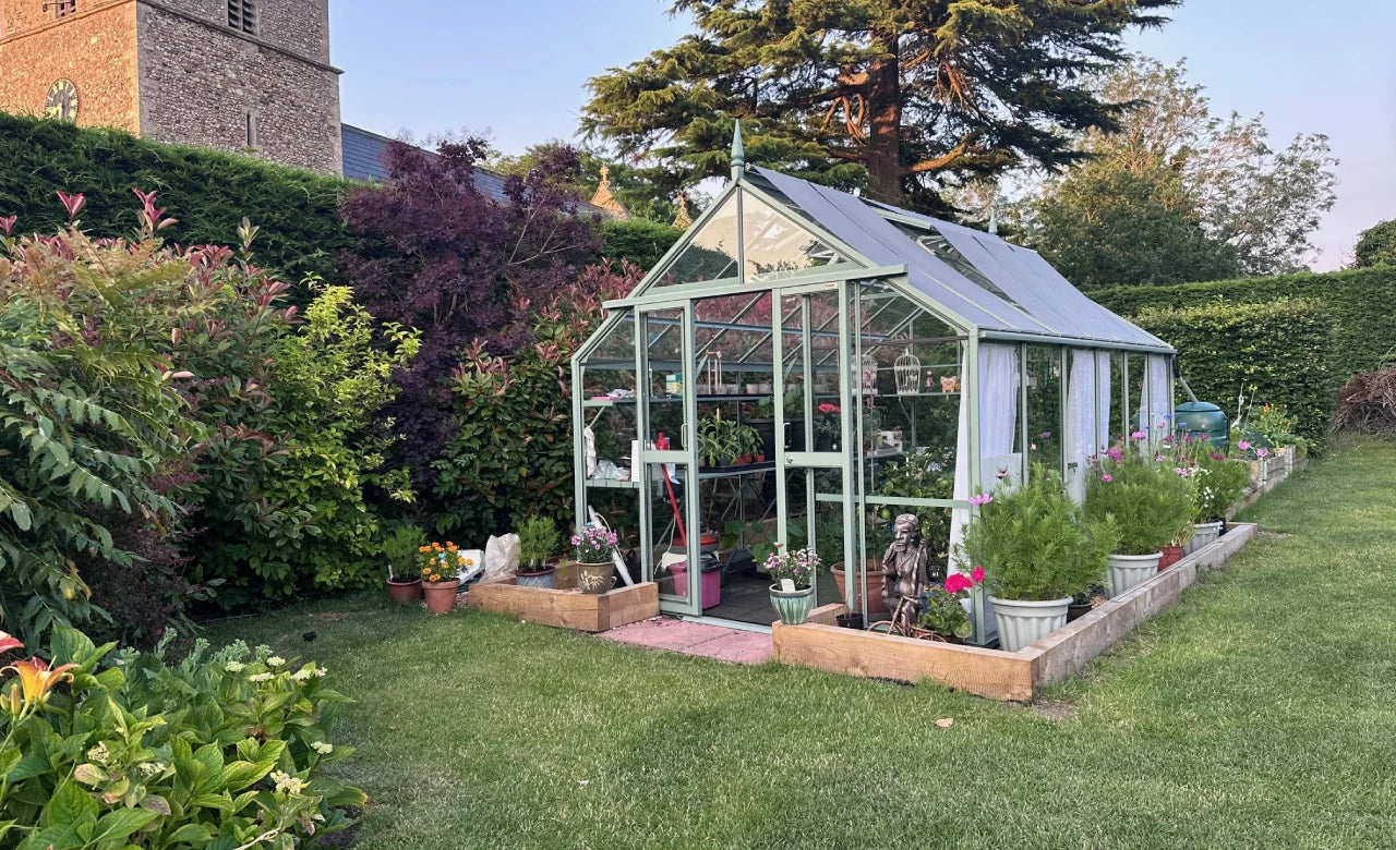 A green, glass-paneled Rhino Greenhouse filled with plants stands on neatly mowed grass, surrounded by lush hedges and colorful shrubs, beside a brick building with a clock tower.