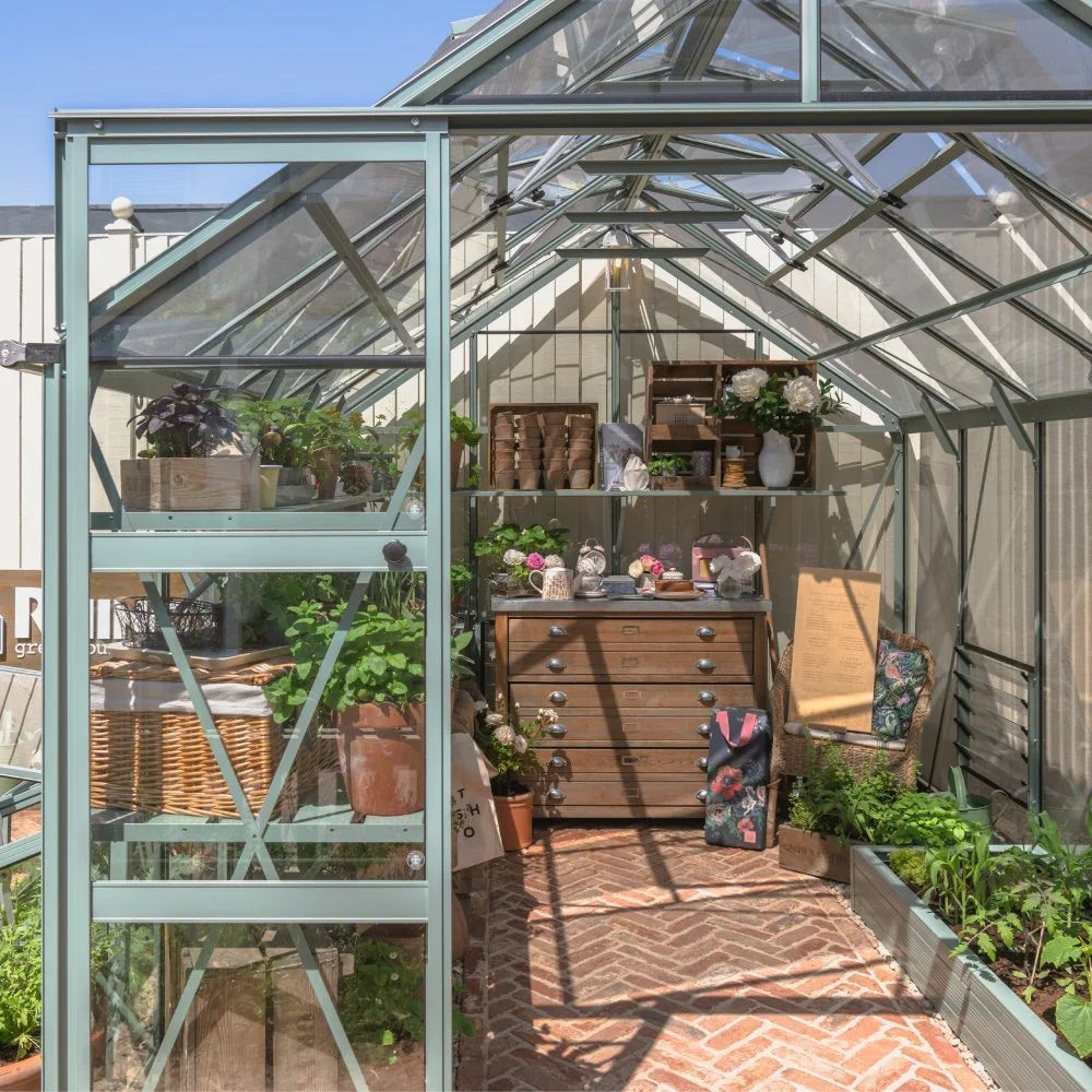 Rhino Greenhouse interior filled with potted plants on shelves. A wooden chest of drawers holds gardening tools, and a wicker basket is nearby. Sunlight streams through glass panels, illuminating the space.
