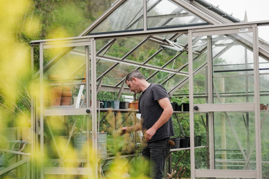 A person tends plants inside a glass Rhino Greenhouse filled with potted greenery and gardening tools. Yellow flowers and lush greenery surround the structure. A small sign reads Rhino.