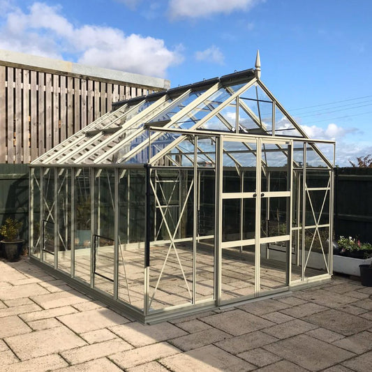 A glass Rhino Greenhouse stands on a paved patio, surrounded by a wooden fence and under a clear blue sky with clouds.
