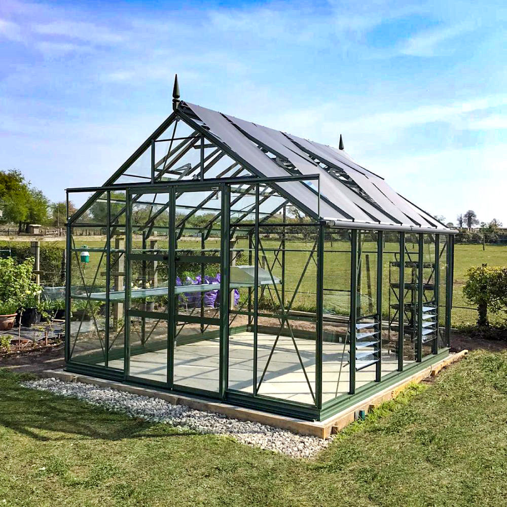 A glass Rhino Greenhouse stands in a grassy field. It features a robust metal frame and multiple shelves, with open skies and trees in the background, creating a serene outdoor setting.