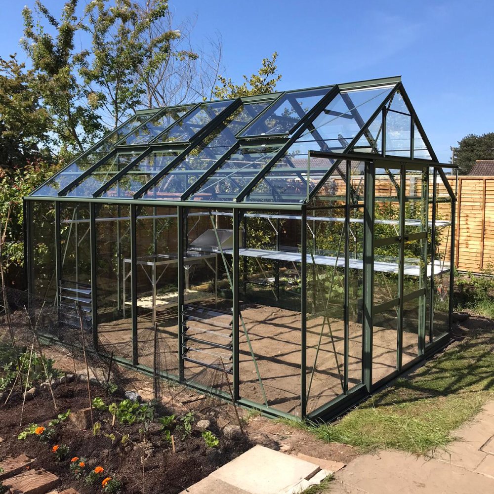 A large glass and metal Rhino Greenhouse stands in a garden, surrounded by plants and trees, under a clear blue sky.