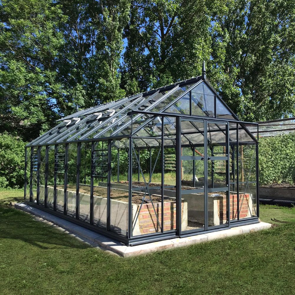 A glass Rhino Greenhouse with a dark metal frame stands on a concrete base, surrounded by grass and lush trees, allowing natural sunlight to filter through its structure.