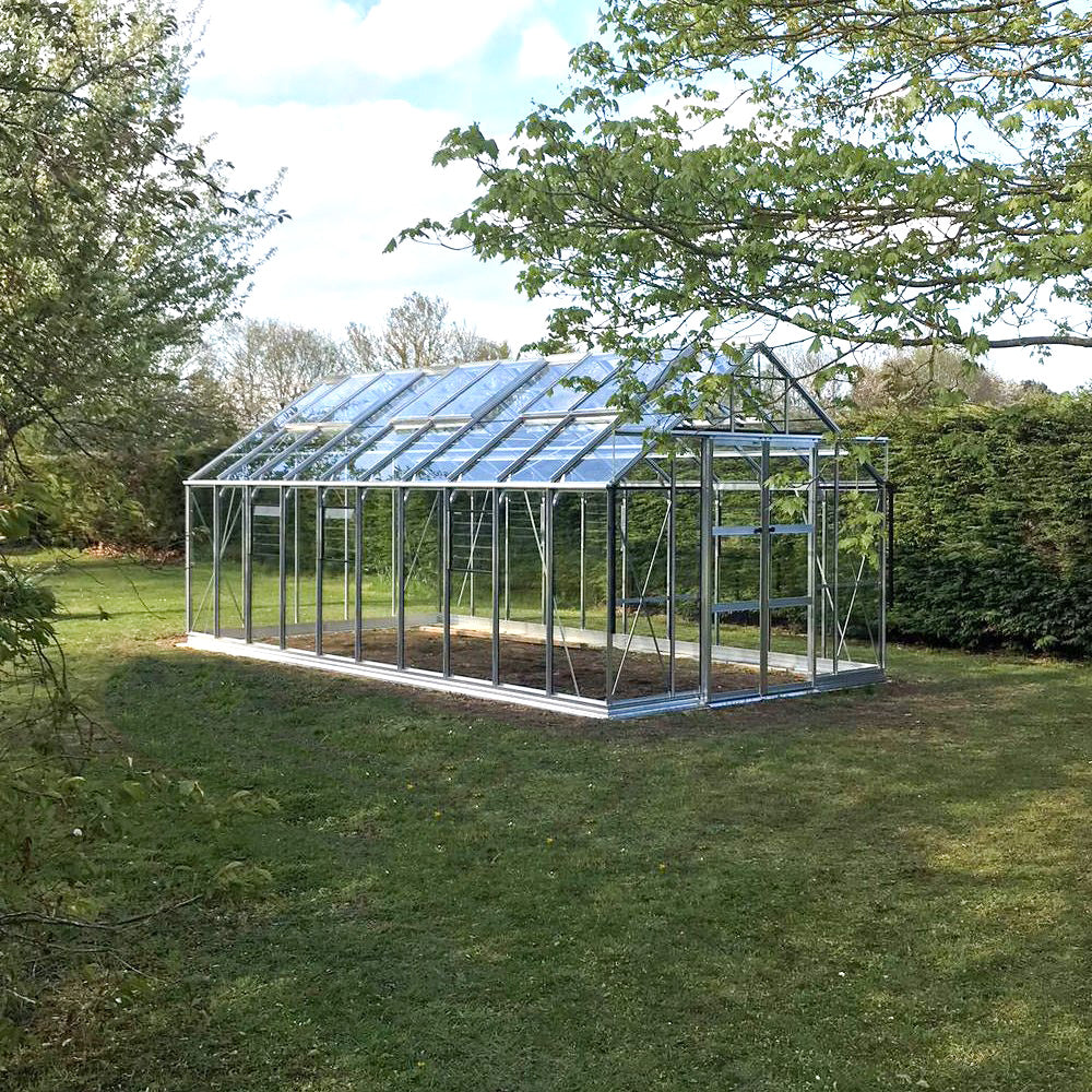 A large glass Rhino Greenhouse stands on a grassy lawn, amidst a backdrop of trees and bushes, under a partly cloudy sky.