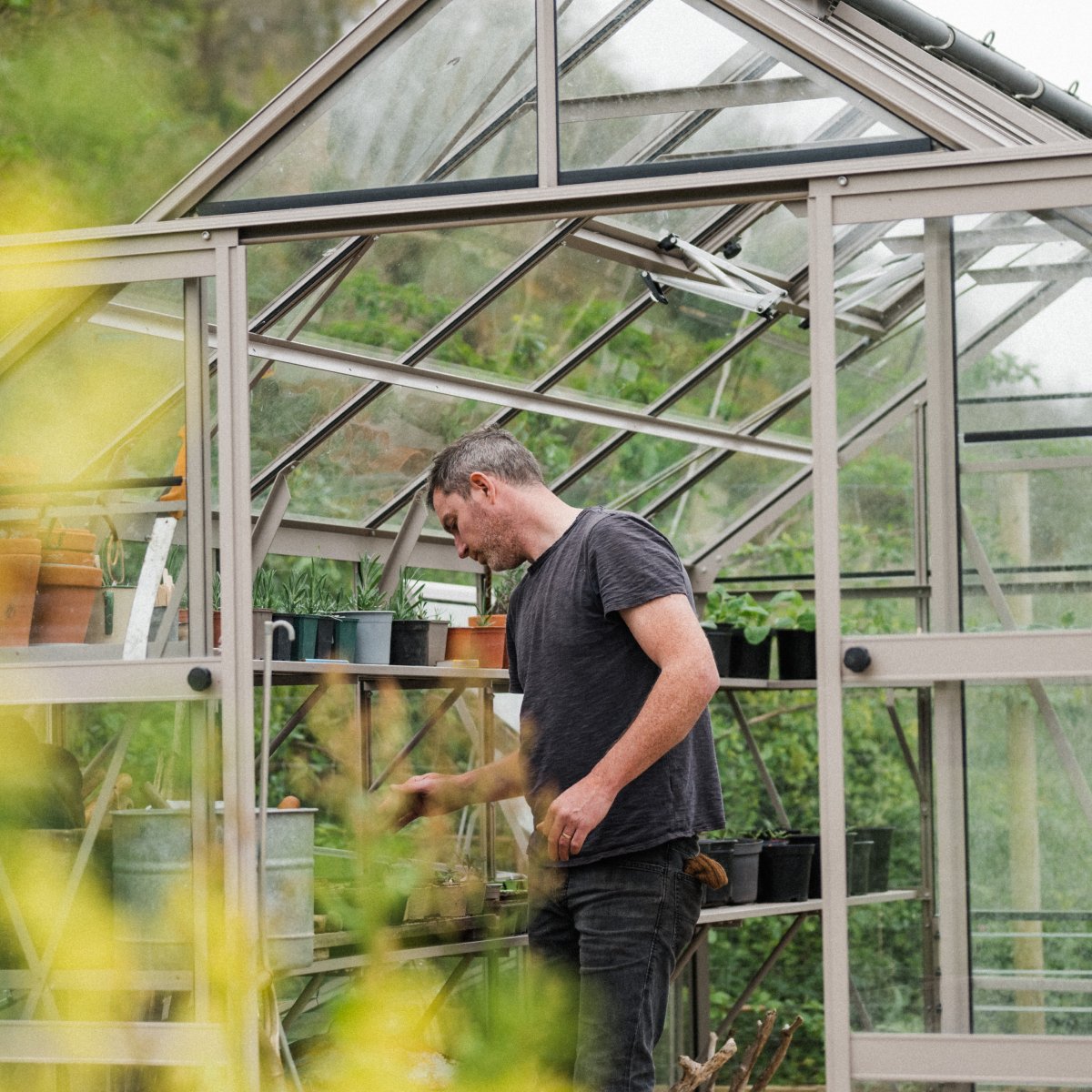 A person tends to plants on shelves inside a glass-paneled Rhino Greenhouse surrounded by lush greenery. Pots and gardening tools are visible in the neatly organized space.