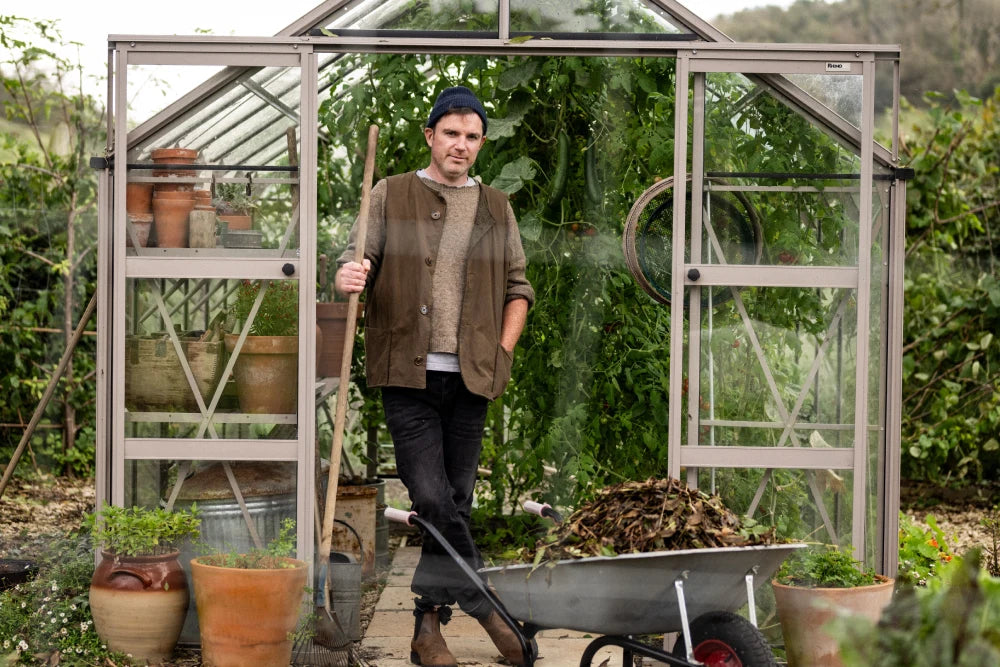 A person holding a rake stands in a glass Rhino Greenhouse filled with plants, surrounded by gardening pots and a wheelbarrow with leaves. Text reads Rhino on the Rhino Greenhouse frame.