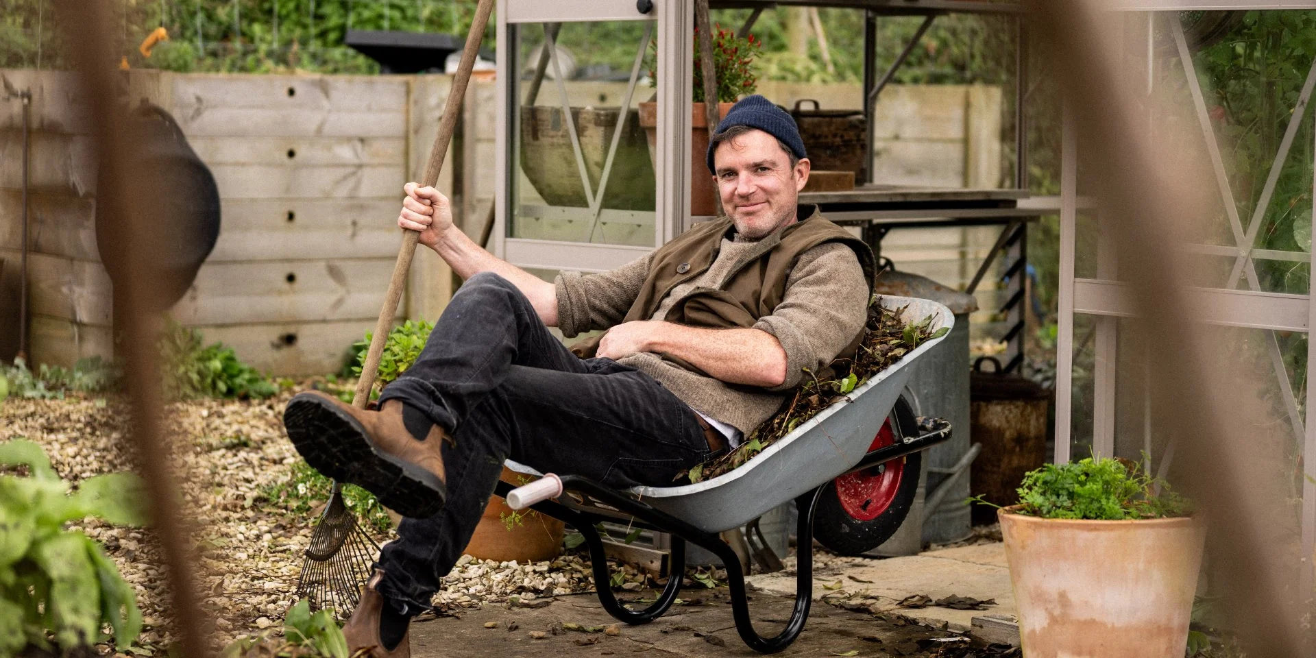 Man relaxing in a wheelbarrow, holding a gardening tool, beside a Rhino Greenhouse with wooden fencing and plants in the background.