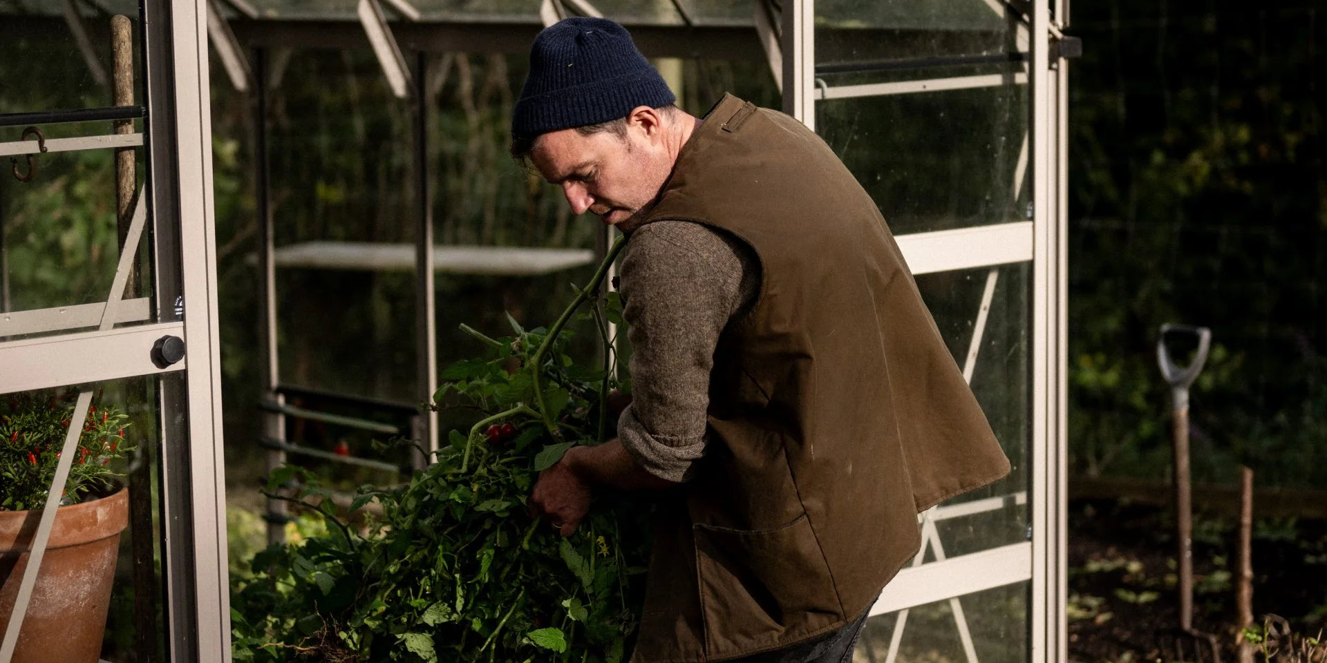 Gardener pruning plants inside a Rhino Greenhouse wearing a hat and vest. Nearby, a terracotta pot holds flowers, and a spade is visible in the garden soil outside.