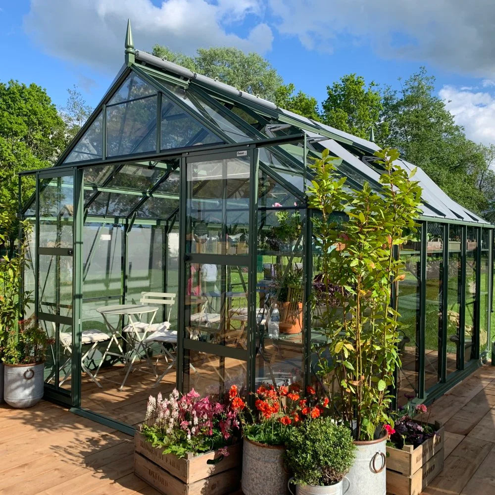 Rhino Greenhouse: A black-framed glass structure housing chairs and plants. Action: Sunlight illuminates the interior. Context: Surrounded by greenery, wooden deck, and colorful potted flowers.