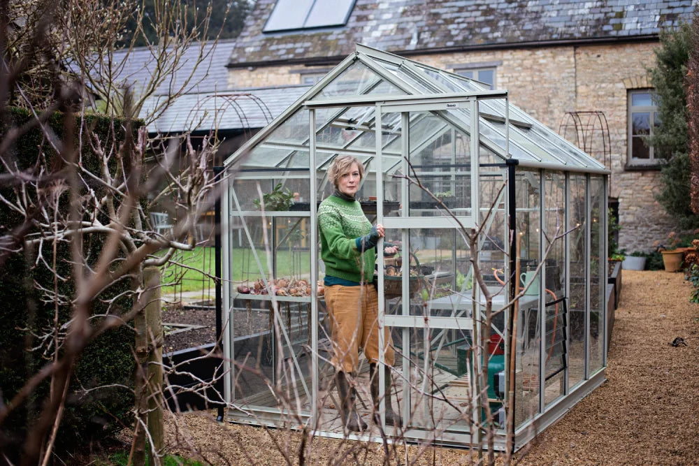 A person stands inside a glass Rhino Greenhouse holding a gardening tool. The Rhino Greenhouse, labeled Rhino, is surrounded by a garden path with bare trees and a stone building nearby.