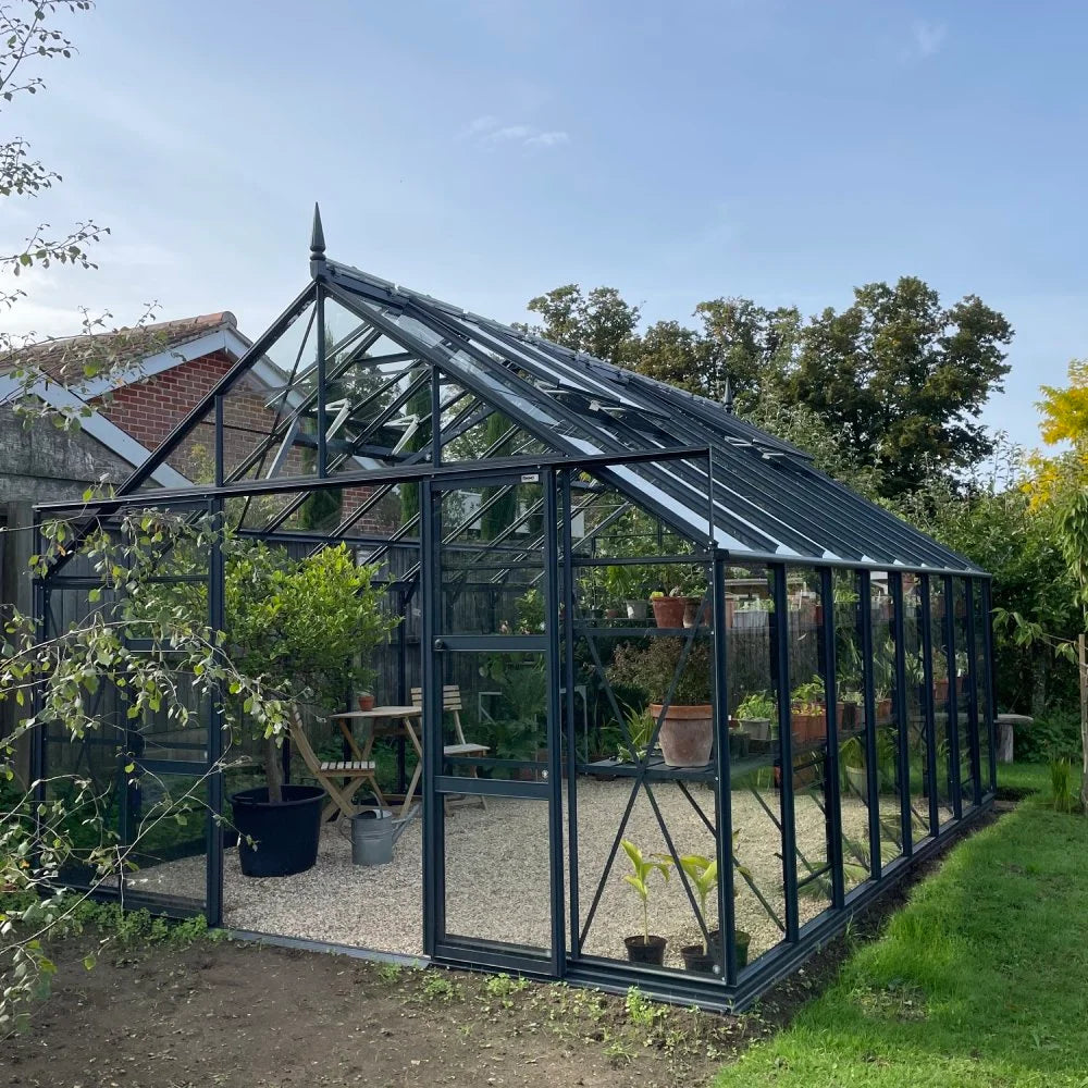 A large, glass Rhino Greenhouse with a dark metal frame houses various plants in pots and hanging shelves. It stands on gravel, surrounded by grass and trees, adjacent to a brick building.