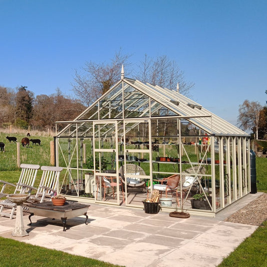 A Rhino Greenhouse stands on a tiled patio, housing plants and garden furniture. Surrounding the structure is a grassy field with grazing cows and distant trees under a clear blue sky.