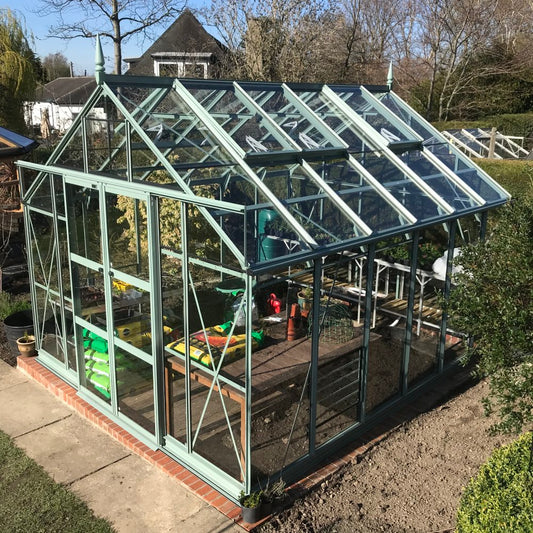 A glass-paneled Rhino Greenhouse stands on a brick base, containing gardening supplies like soil bags and pots. It is situated in a garden with trees and a house visible in the background.