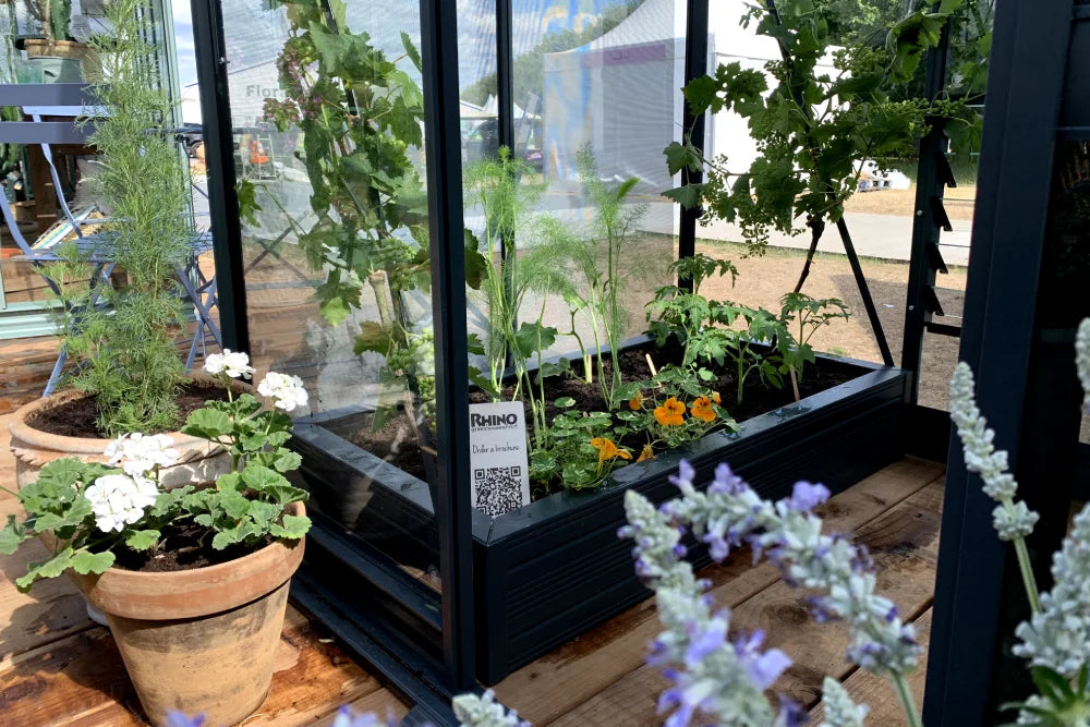 Rhino Greenhouse containing various plants inside, including flowers and vegetables, displayed outdoors on a wooden platform. Nearby, potted plants and a QR code with Rhino text are visible.