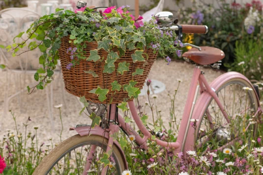 A pink bicycle with a wicker basket overflows with greenery and pink flowers, parked amid a garden setting with wildflowers and outdoor furniture in the background.