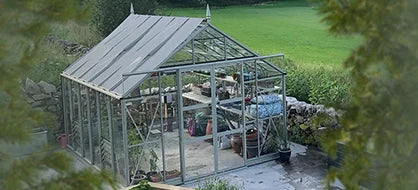 A glass Rhino Greenhouse stands in a garden, housing various plants and tools. It's surrounded by stone walls and lush greenery, with fields and trees in the background.