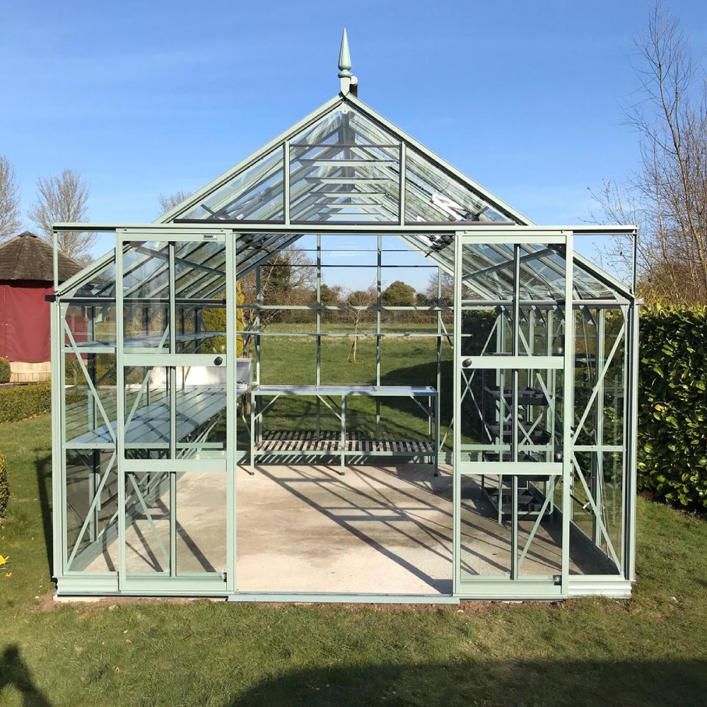 Rhino Greenhouse with green metal frame and glass panels stands on grass. Inside, shelves and tables are arranged. Surrounding are trees and a blue sky, creating a serene garden atmosphere.