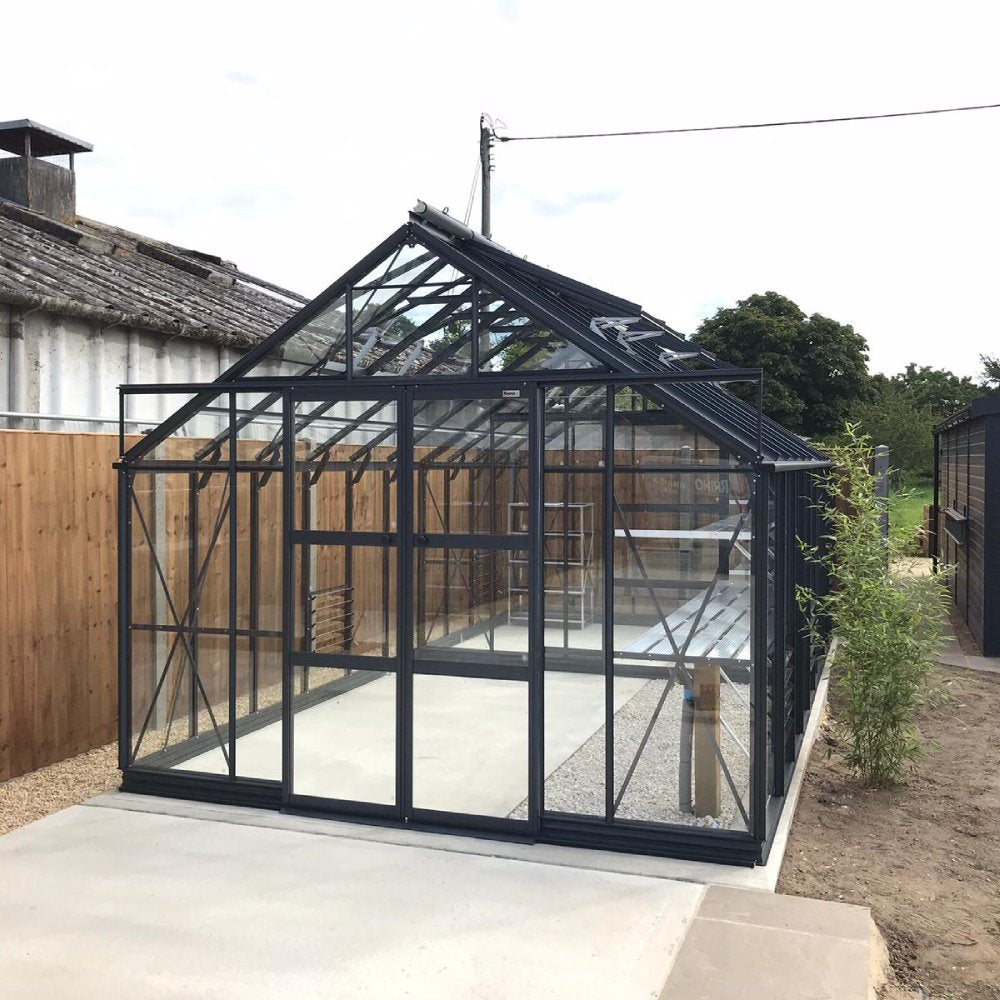 A glass and metal Rhino Greenhouse stands on a concrete base, surrounded by a wooden fence and adjacent structures under a partly cloudy sky.